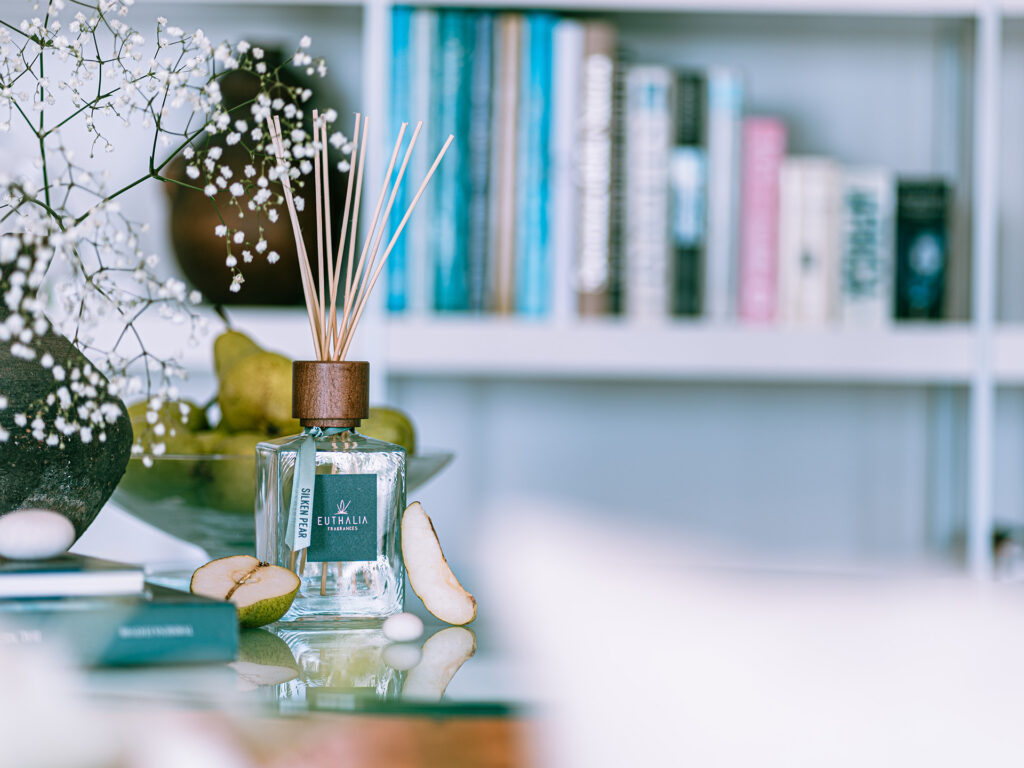 Diffusore di fragranza ambiente Silken Pear di Euthalia Fragrances con bastoncini in legno, esposto su un tavolo con pere fresche e fiori bianchi, in un elegante ambiente domestico con libreria sullo sfondo. Nuove fragranze per la tua casa. New fragrances for your home.