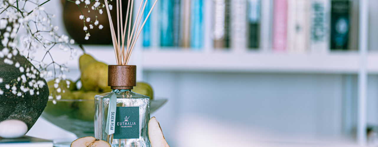 Diffusore di fragranza ambiente Silken Pear di Euthalia Fragrances con bastoncini in legno, esposto su un tavolo con pere fresche e fiori bianchi, in un elegante ambiente domestico con libreria sullo sfondo. Nuove fragranze per la tua casa. New fragrances for your home.