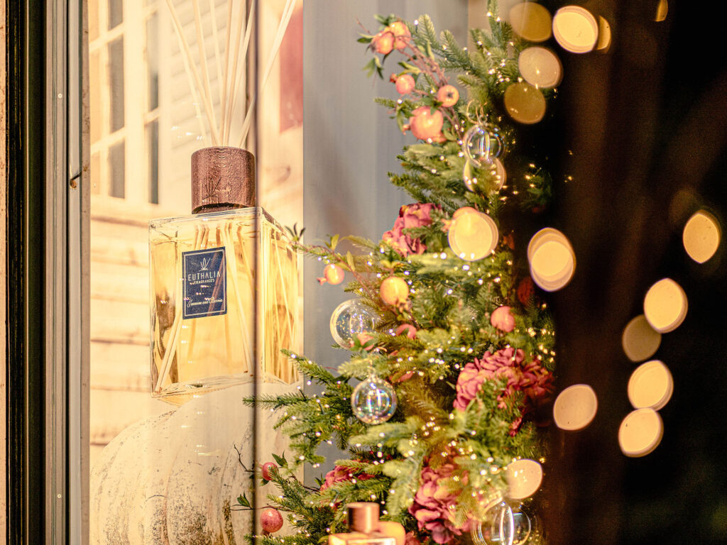 Diffusore Euthalia Fragrances in vetrina natalizia con albero decorato, luci calde e dettagli floreali per creare atmosfere eleganti. Euthalia Fragrances reed diffuser in a Christmas window display with decorated tree, warm lights and floral details, creating refined atmospheres