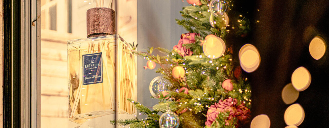 Diffusore Euthalia Fragrances in vetrina natalizia con albero decorato, luci calde e dettagli floreali per creare atmosfere eleganti. Euthalia Fragrances reed diffuser in a Christmas window display with decorated tree, warm lights and floral details, creating refined atmospheres