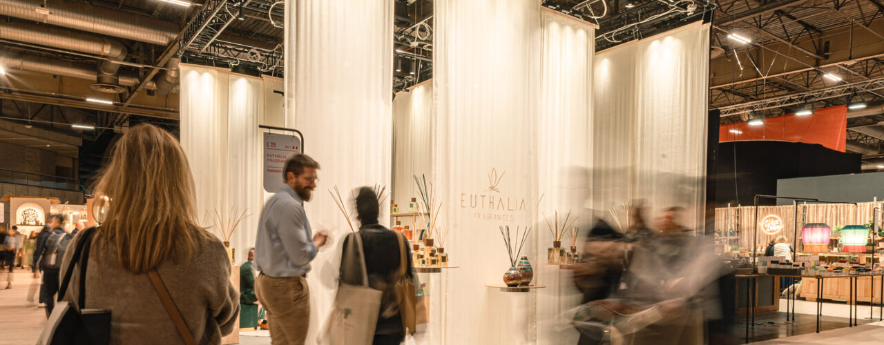 Inizio 2026: stand Euthalia Fragrances a Maison&Objet con tende bianche, esposizione di diffusori e visitatori in fiera. Start of 2026: Euthalia Fragrances booth at Maison&Objet with white drapes, diffuser display and visitors walking through the trade show.