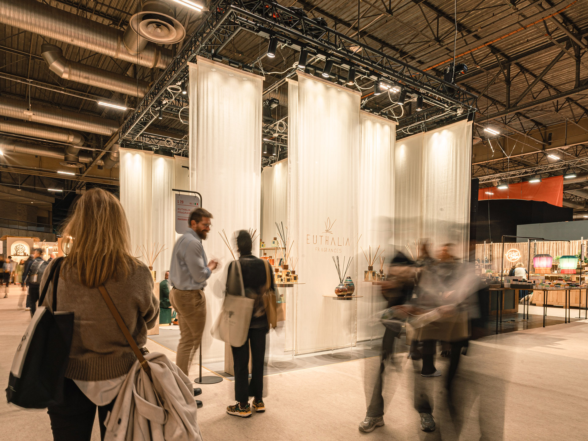 Inizio 2026: stand Euthalia Fragrances a Maison&Objet con tende bianche, esposizione di diffusori e visitatori in fiera. Start of 2026: Euthalia Fragrances booth at Maison&Objet with white drapes, diffuser display and visitors walking through the trade show.