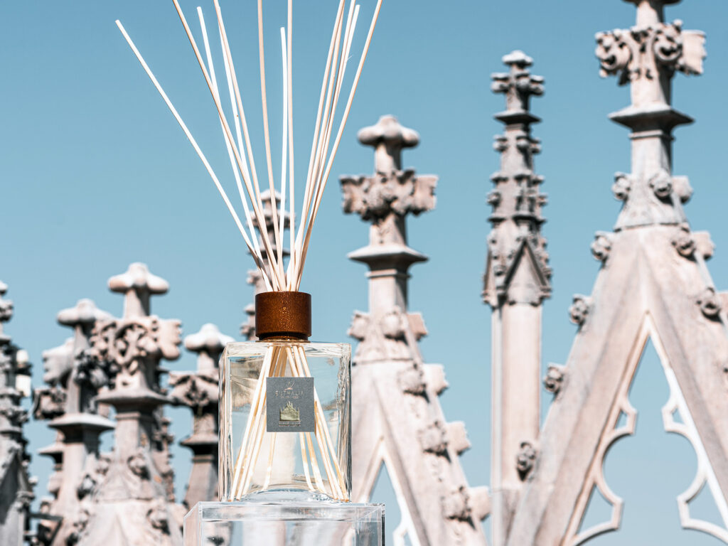Diffusore Duomo Milano sul Duomo di Milano, tra guglie gotiche e cielo azzurro, ispirato al profumo della neve e all’atmosfera di Milano Cortina 2026. Duomo Milano reed diffuser on the Duomo rooftop in Milan, among Gothic spires and blue sky, inspired by the scent of snow and the atmosphere of Milan Cortina 2026.
