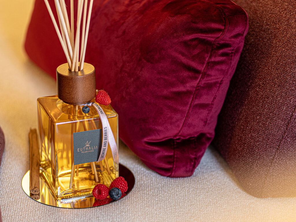 Diffusore per ambiente Euthalia Fragrances in una scena romantica con cuscini bordeaux, frutti rossi e atmosfera calda ed elegante. Euthalia Fragrances reed diffuser in a romantic setting with burgundy cushions, red berries, and a warm elegant atmosphere.