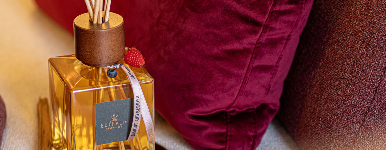 Diffusore per ambiente Euthalia Fragrances in una scena romantica con cuscini bordeaux, frutti rossi e atmosfera calda ed elegante. Euthalia Fragrances reed diffuser in a romantic setting with burgundy cushions, red berries, and a warm elegant atmosphere.