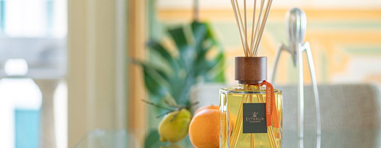 Diffusore per ambiente Plaisir d’Agrume di Euthalia Fragrances su tavolo in vetro con agrumi e cannella, immagine dedicata all’arredo olfattivo di un interno elegante. Euthalia Fragrances Plaisir d’Agrume reed diffuser on a glass table with citrus fruits and cinnamon, an elegant interior image inspired by olfactory decor