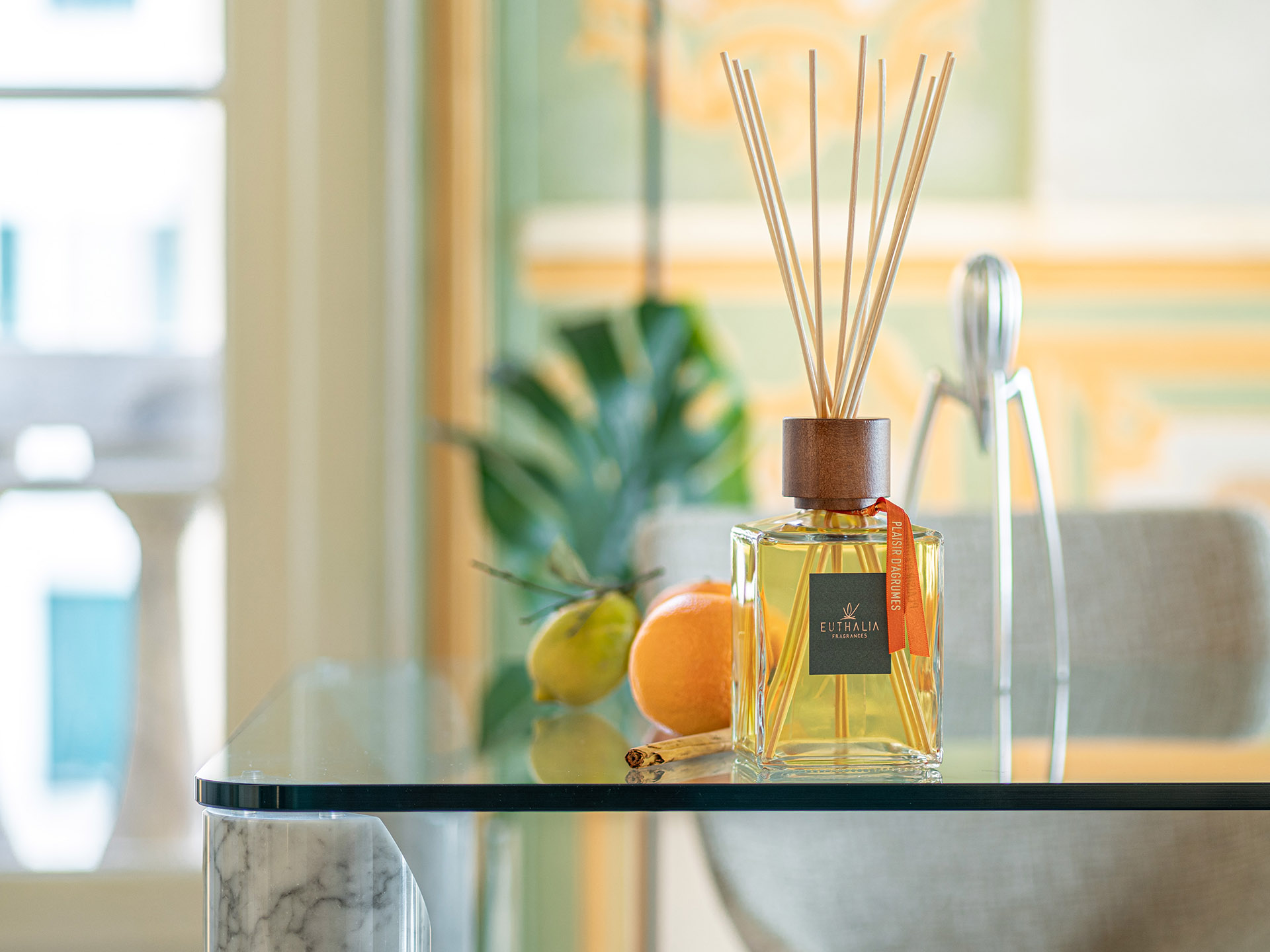 Diffusore per ambiente Plaisir d’Agrume di Euthalia Fragrances su tavolo in vetro con agrumi e cannella, immagine dedicata all’arredo olfattivo di un interno elegante. Euthalia Fragrances Plaisir d’Agrume reed diffuser on a glass table with citrus fruits and cinnamon, an elegant interior image inspired by olfactory decor